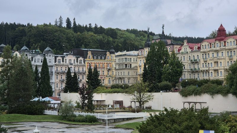 Historische Kurhäuser in Marienbad vor einem bewaldeten Hang, im Vordergrund die Parkanlagen
