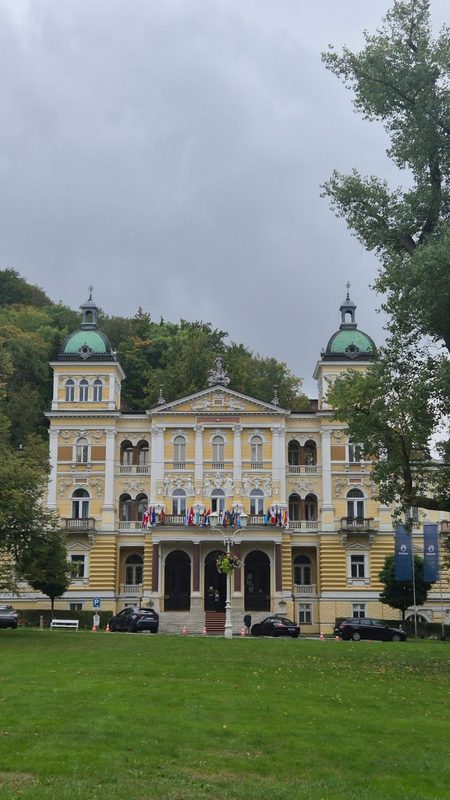 Gelbe Fassade des Hotels Nové Lázně in Marienbad mit zwei grünen Kuppeltürmen und Fahnen über dem Eingang