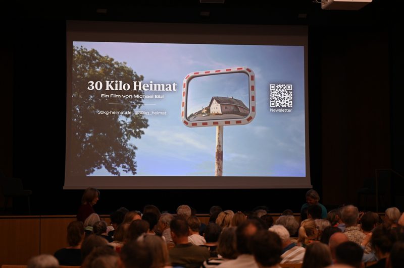 Leinwand mit dem Titel „30 Kilo Heimat“ und einem Verkehrsspiegel vor dem voll besetzten Saal