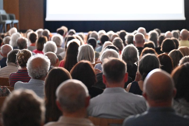 Publikum im voll besetzten Saal von hinten, vor der hellen Leinwand